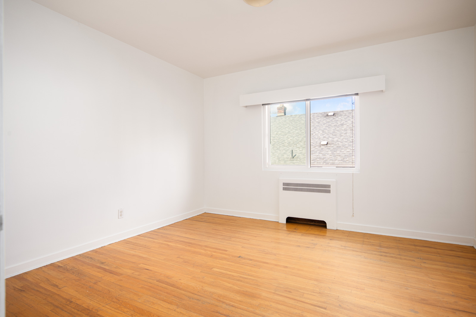Empty room with white walls, a wooden floor, a window with partial outdoor view, and a wall-mounted heater below the window.