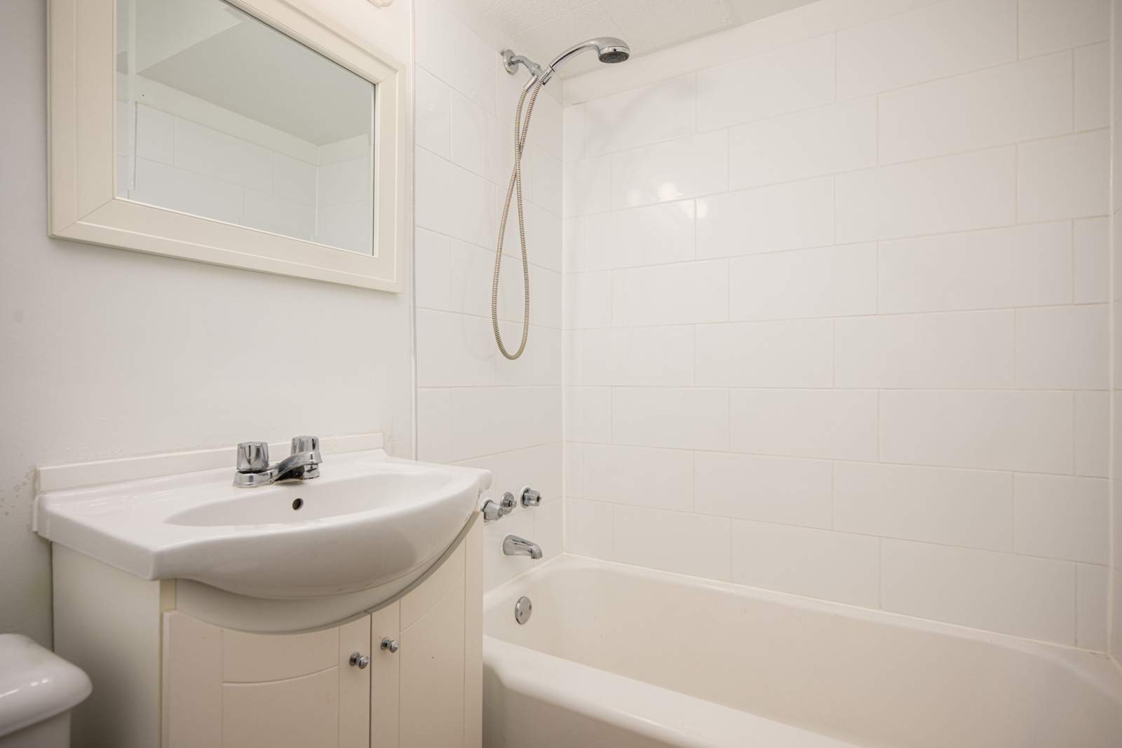 A white bathroom with a wall-mounted mirror, sink with a cabinet, and a bathtub featuring a handheld showerhead.