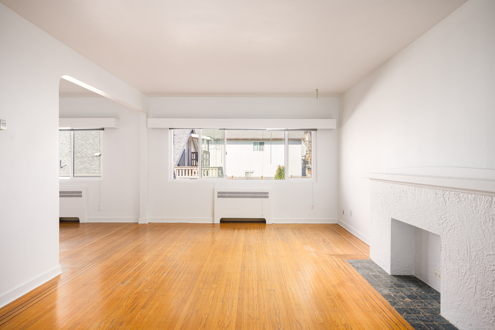 Unfurnished living room with white walls, hardwood floors, large windows, and a white textured fireplace with a tiled hearth.
