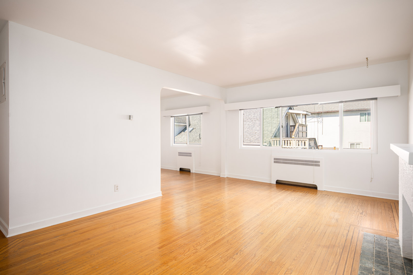 Empty, sunlit living room with hardwood floors, white walls, and large windows overlooking neighboring buildings.