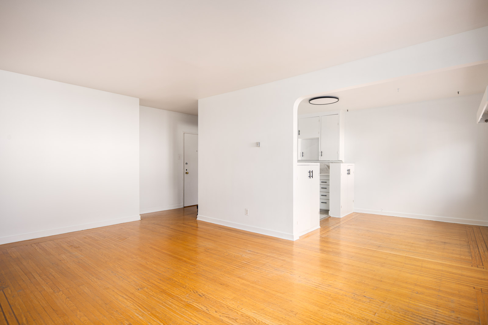 Empty room with light wood floors, white walls, and an open archway leading to a small kitchen area.