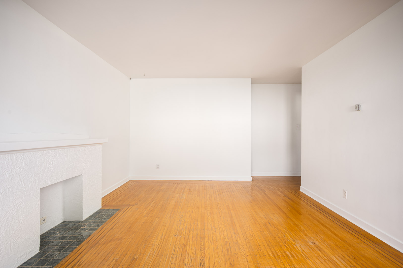 Empty room with white walls, light wood flooring, a small white fireplace on the left, and an open doorway in the back right.