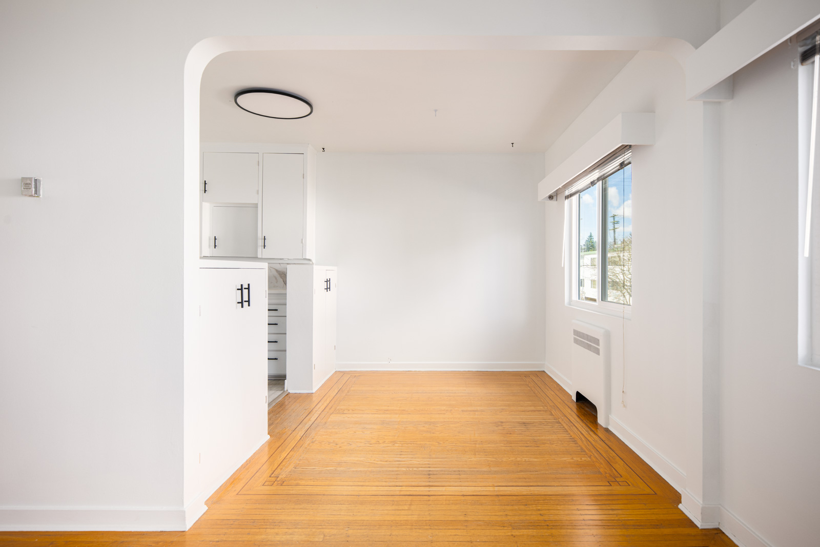Bright, empty room with hardwood floors, white walls, a window with blinds, and a partial view of an adjacent kitchen area.