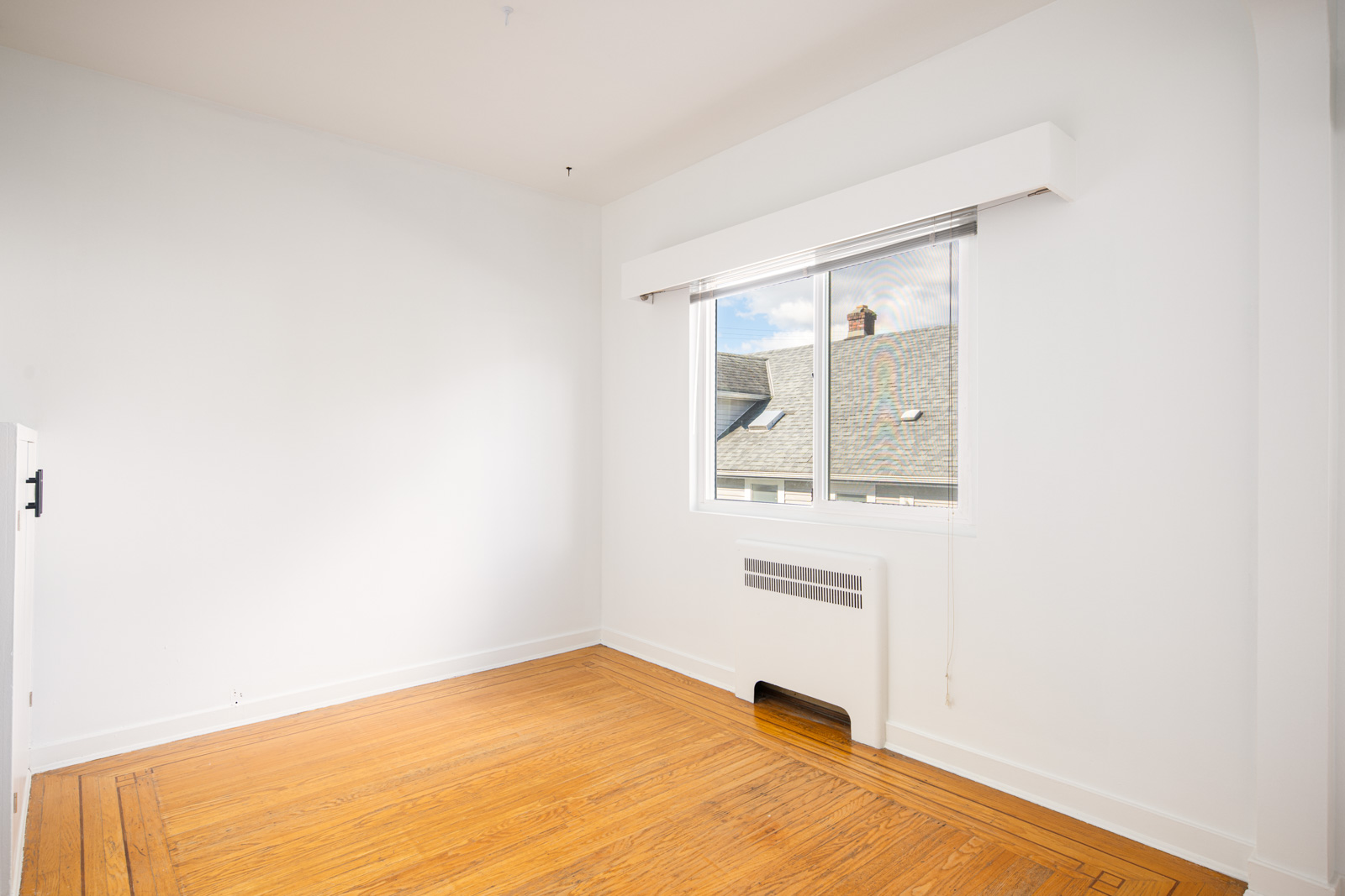 Empty room with white walls, a large window showing rooftops outside, hardwood floors, and a white wall heater below the window.