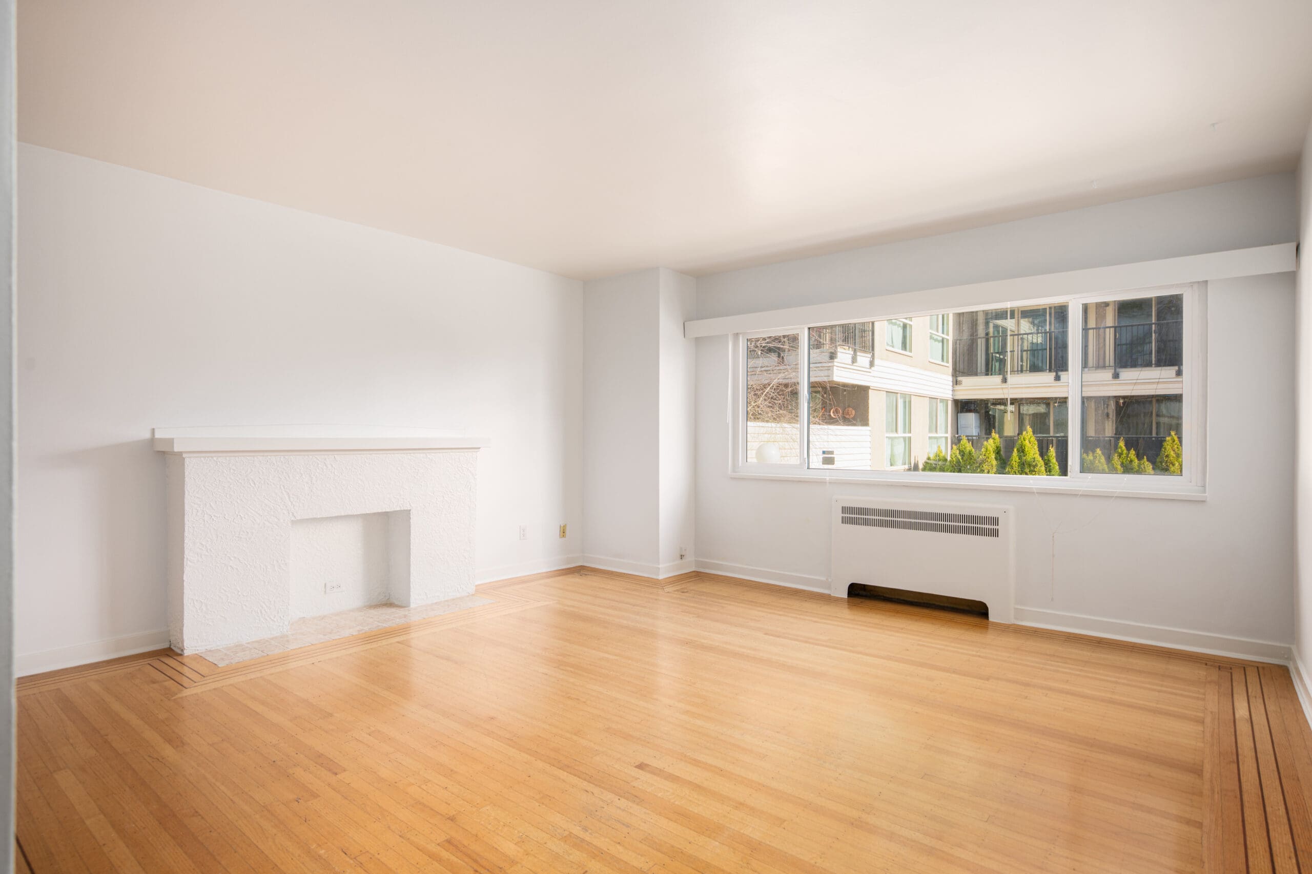 Unfurnished living room with light wood flooring, a white decorative fireplace, large window, and wall heater, overlooking an apartment building exterior.