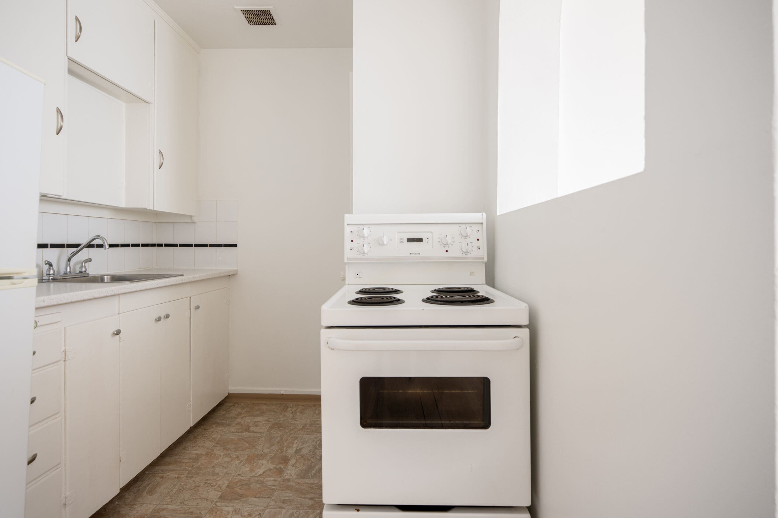A small, white kitchen with a white electric stove, oven, sink, cabinets, and a window letting in natural light.