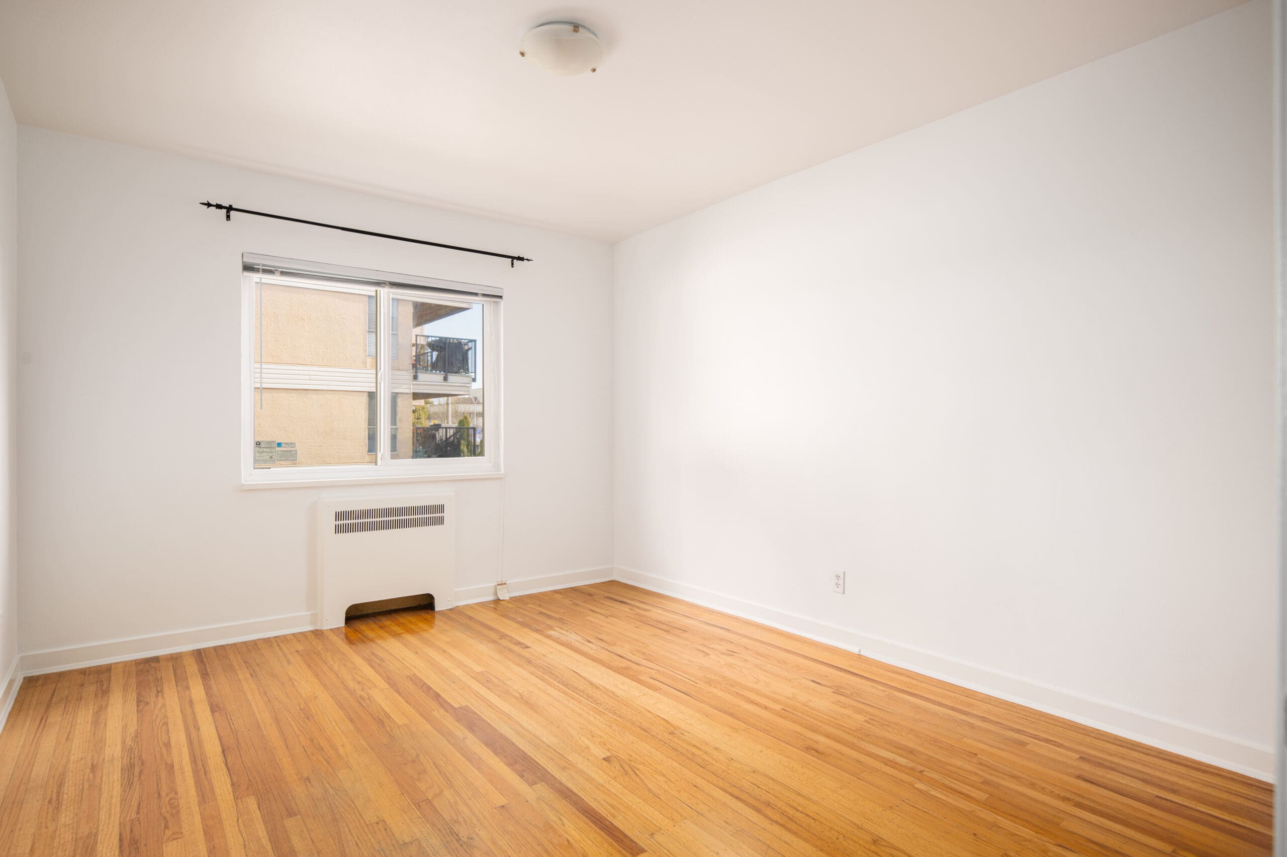 Empty room with white walls, a wooden floor, a window with a black curtain rod, and a radiator beneath the window. Ceiling light fixture is centered above.