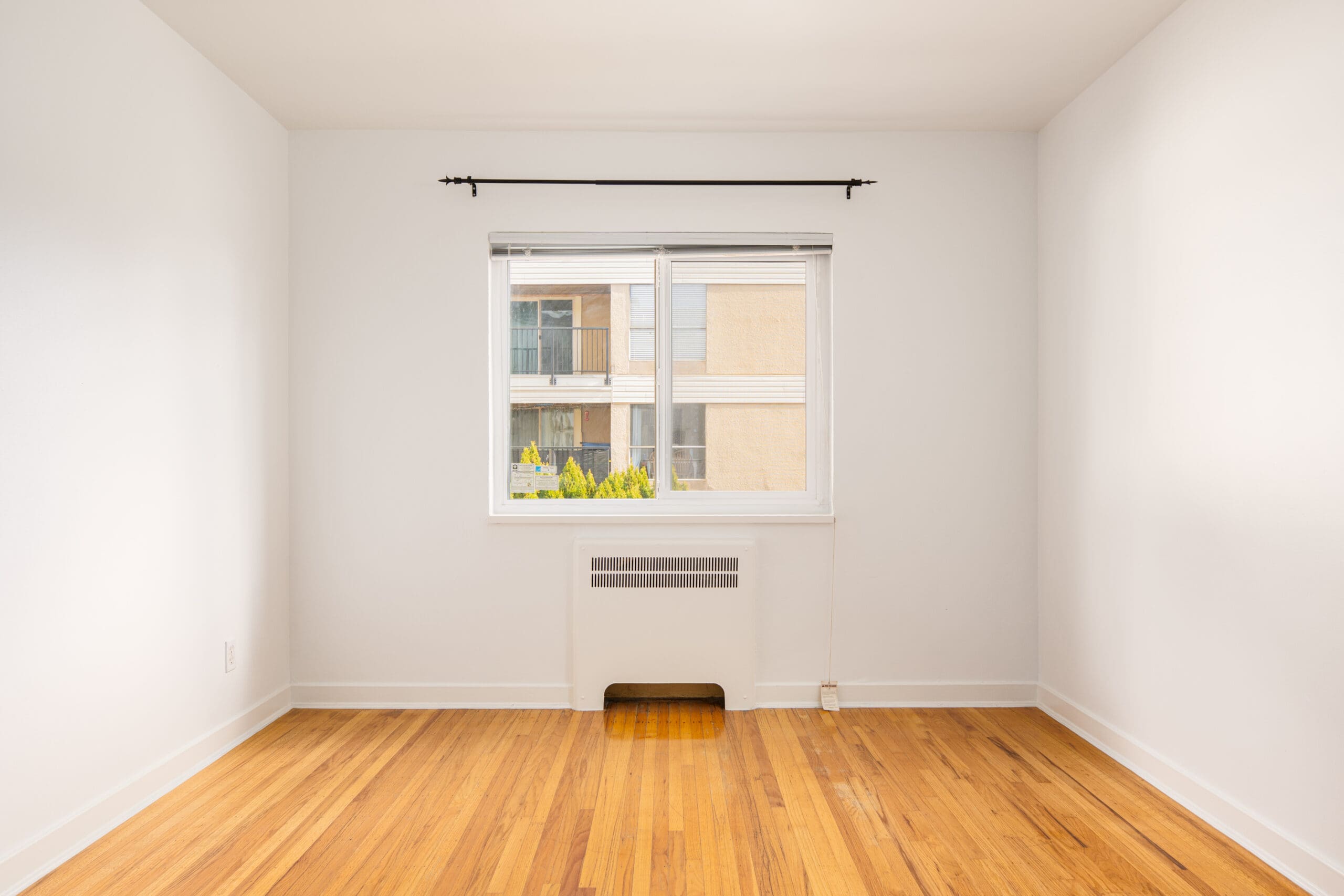Empty room with white walls, a window with a black curtain rod, hardwood floors, and a small wall heater below the window. An apartment building is visible outside.