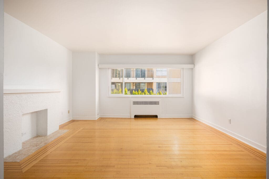 Empty, unfurnished living room with light wood floors, white walls, a fireplace on the left, a large window, and a radiator beneath the window. Apartment buildings visible outside.