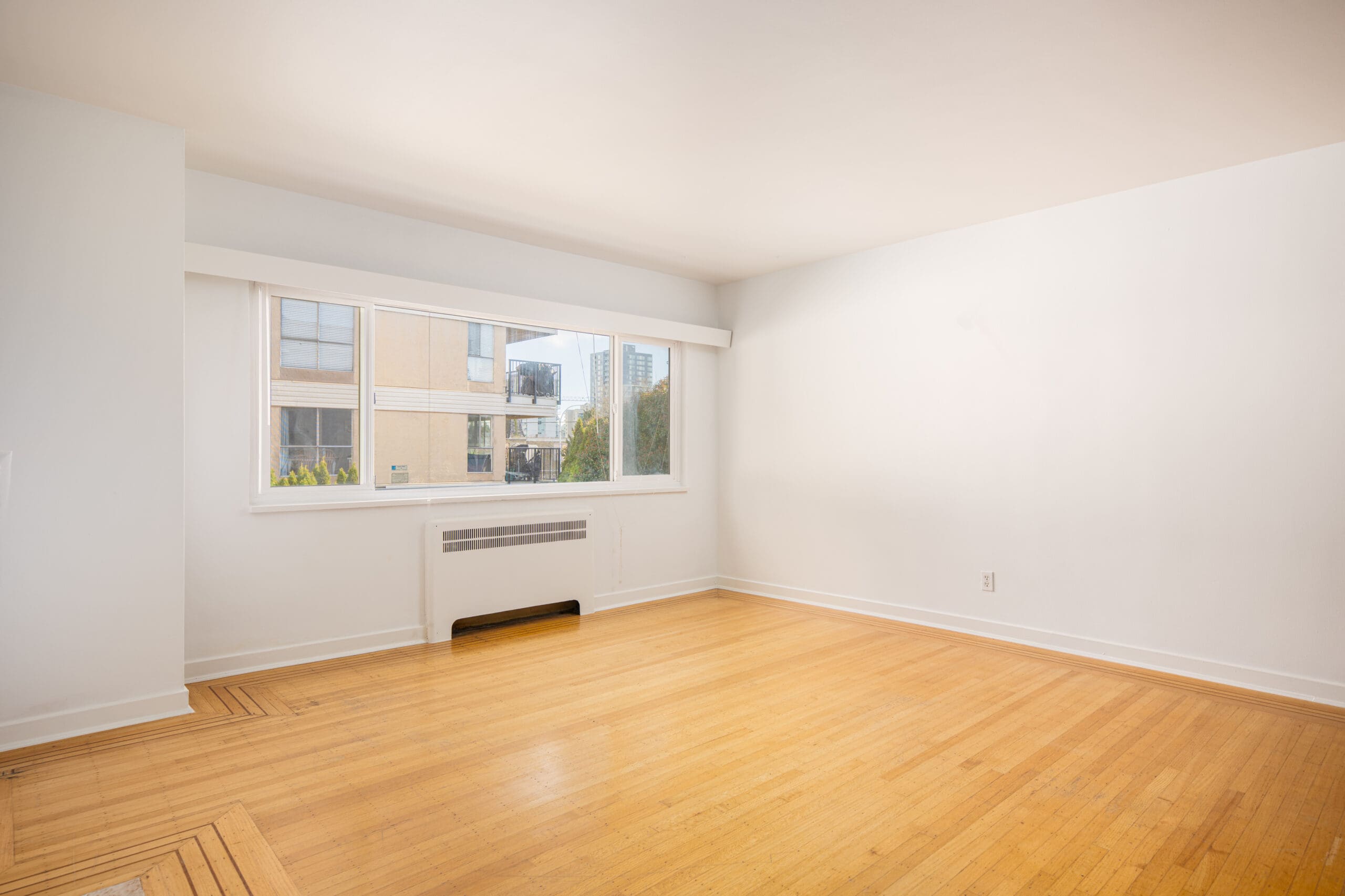 Empty room with light-colored walls, large window, hardwood floors, and a baseboard heater beneath the window. Natural light fills the space.