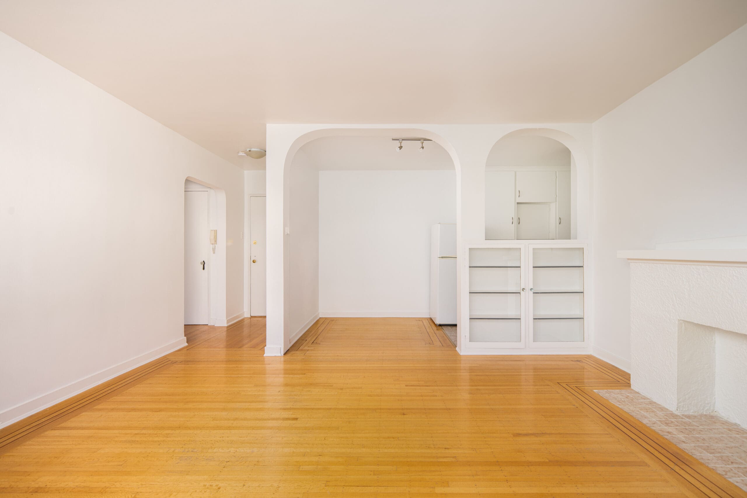 Unfurnished room with light wood floors, white walls, built-in shelves, and an open archway leading to a refrigerator in a small alcove.