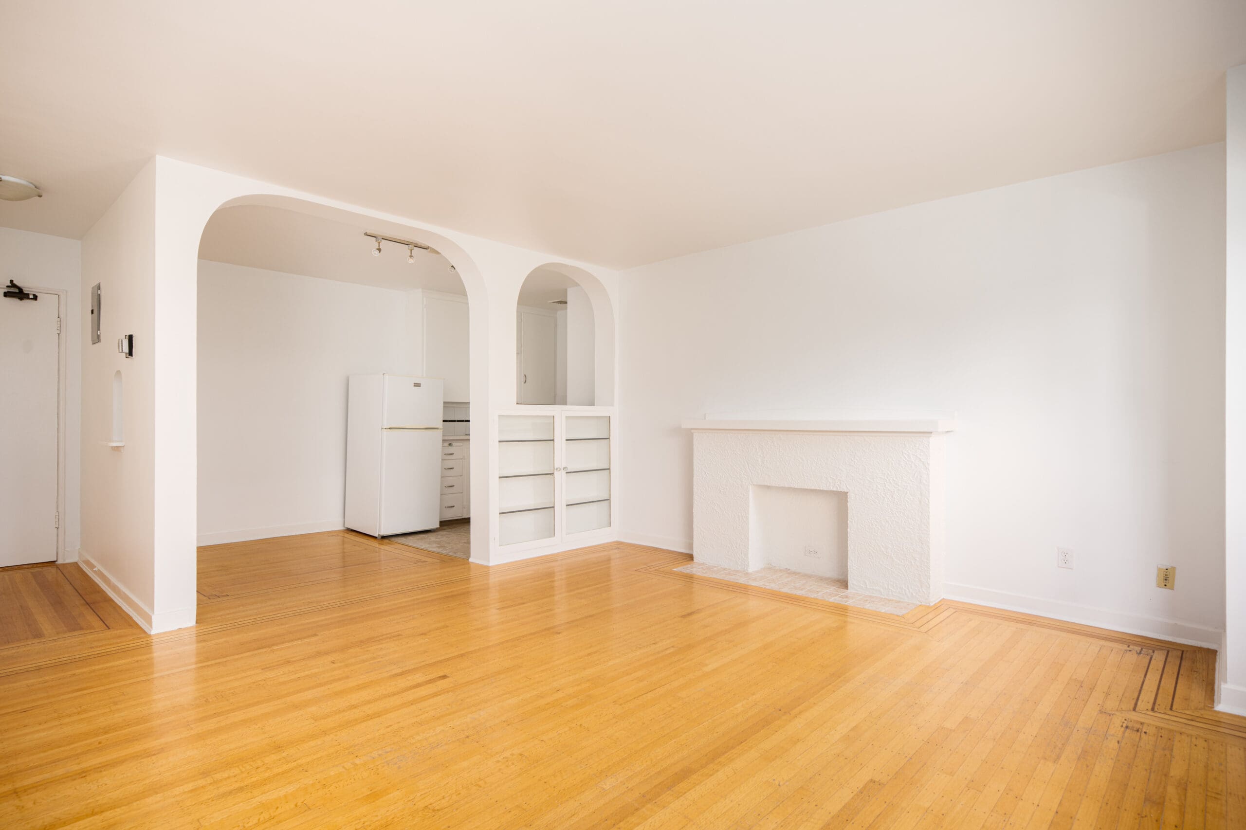 Unfurnished living room with hardwood floor, white walls, a fireplace, built-in shelves, and an open archway leading to a small kitchen with a refrigerator.