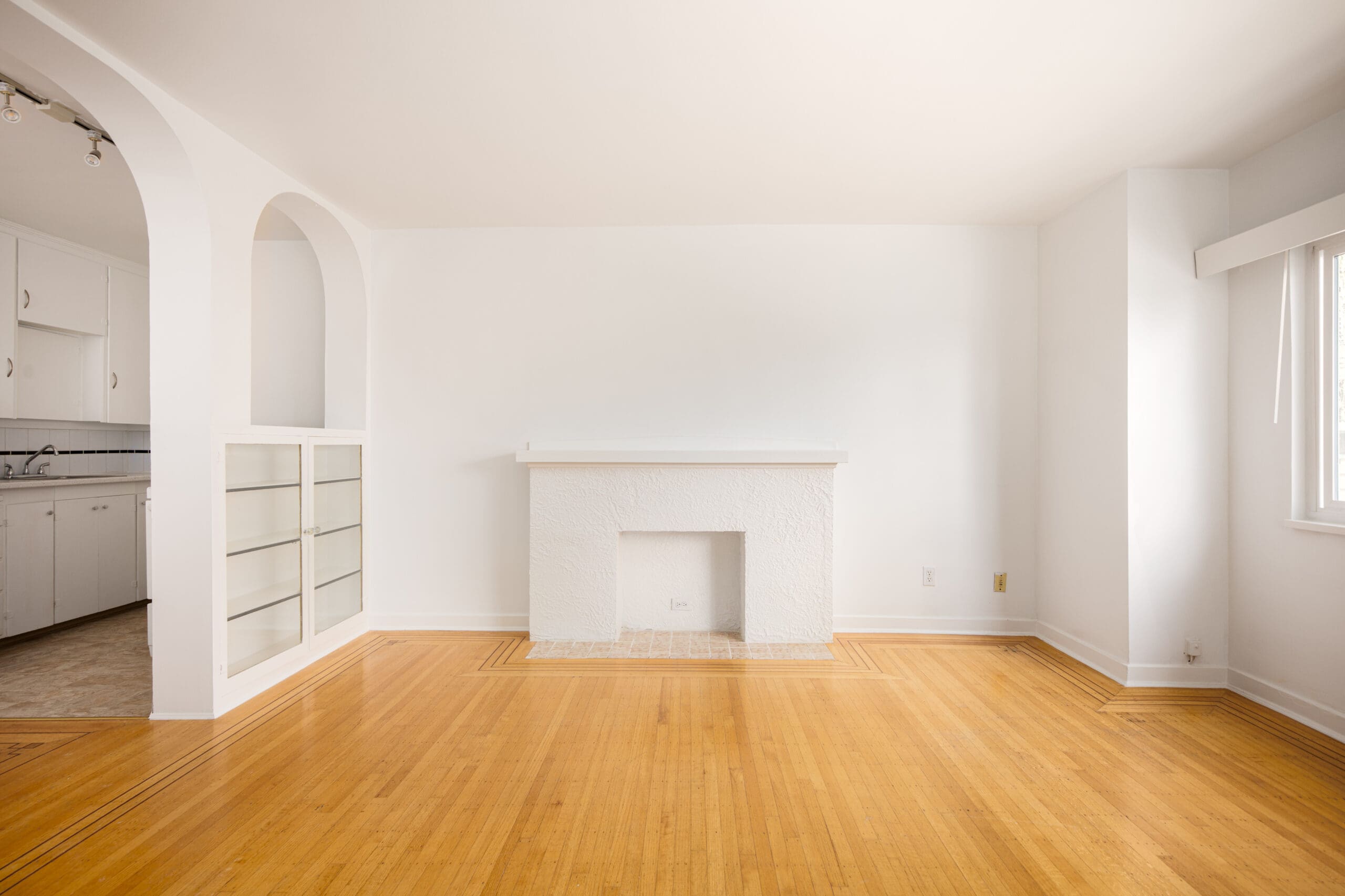 Unfurnished living room with light wood floors, a white fireplace, built-in shelves, and an open doorway leading to a kitchen. Large window on the right lets in natural light.