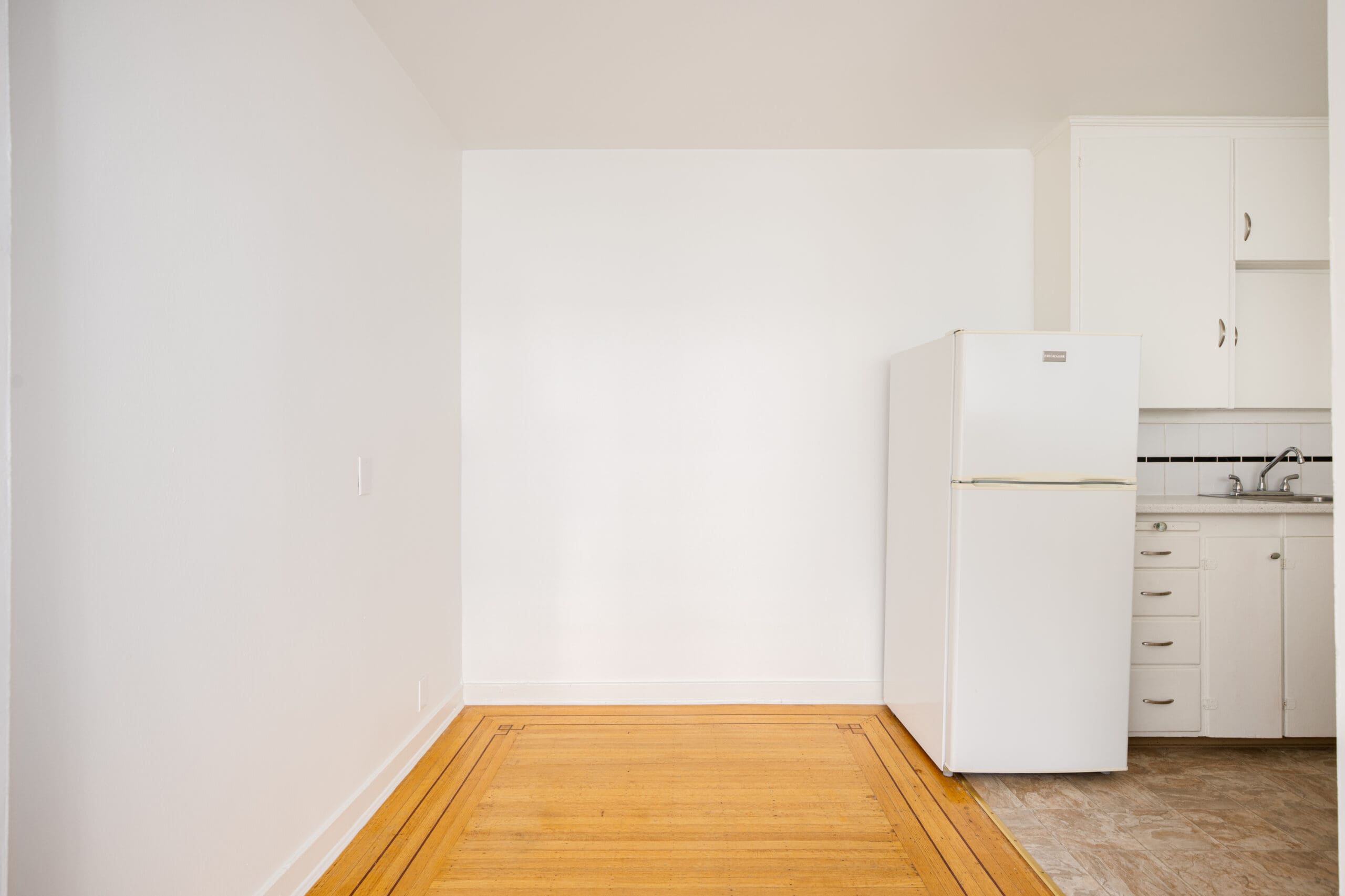 A small kitchen area with a white refrigerator, white cabinets, a sink, and a section of hardwood floor next to tile flooring under bright lighting.