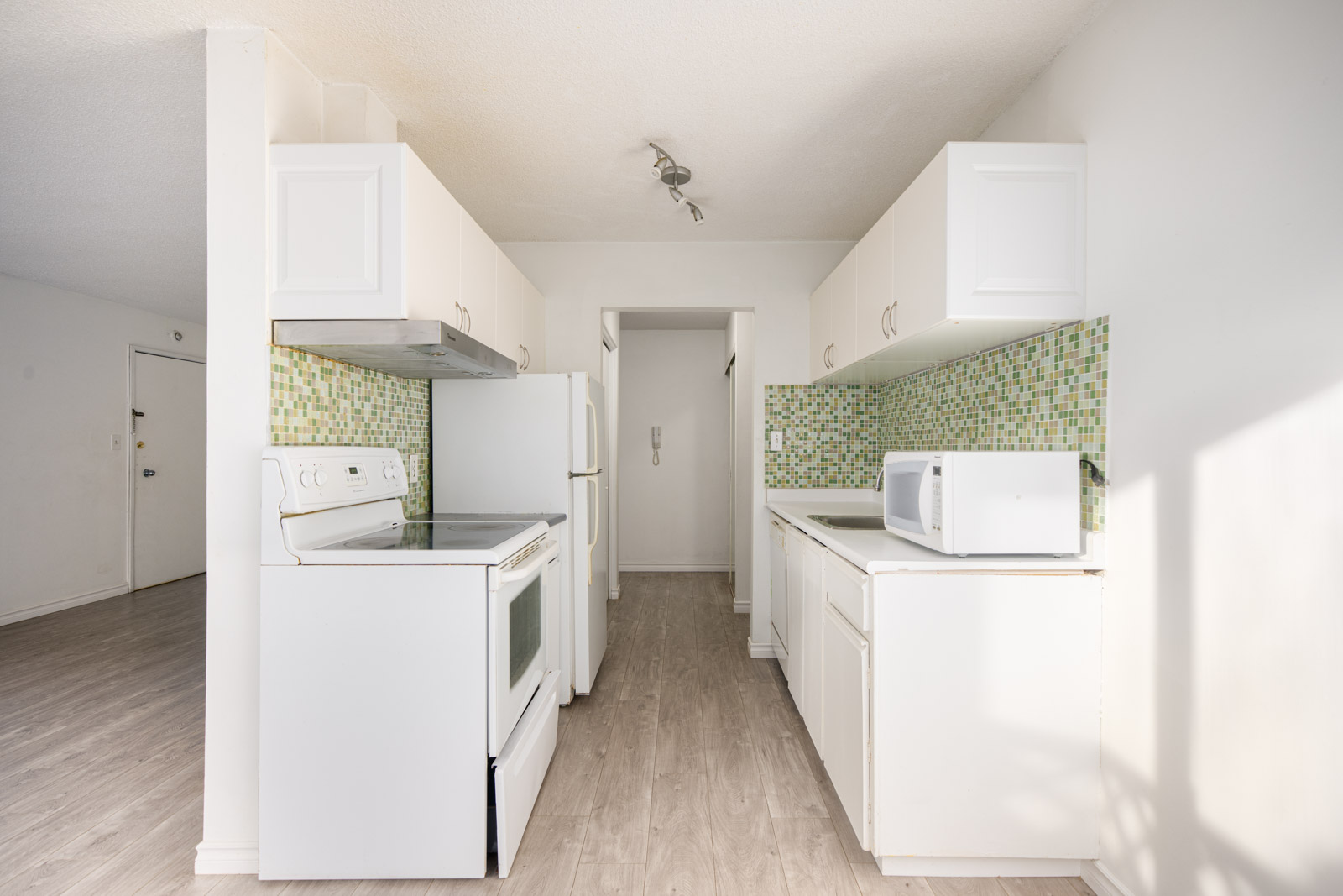 A small kitchen with white cabinets, green mosaic backsplash, white appliances, a microwave, and light wood flooring.