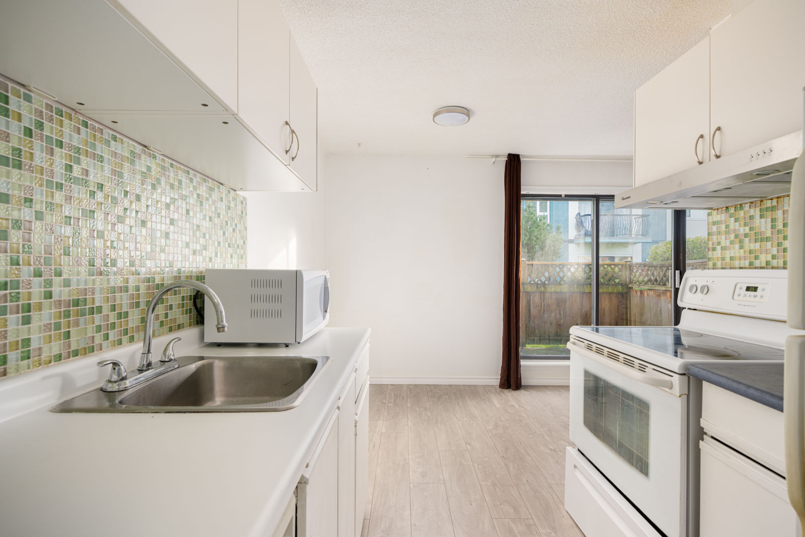 Bright kitchen with white cabinets, green mosaic tile backsplash, white appliances, a microwave on the counter, and a sliding glass door leading to a fenced backyard.