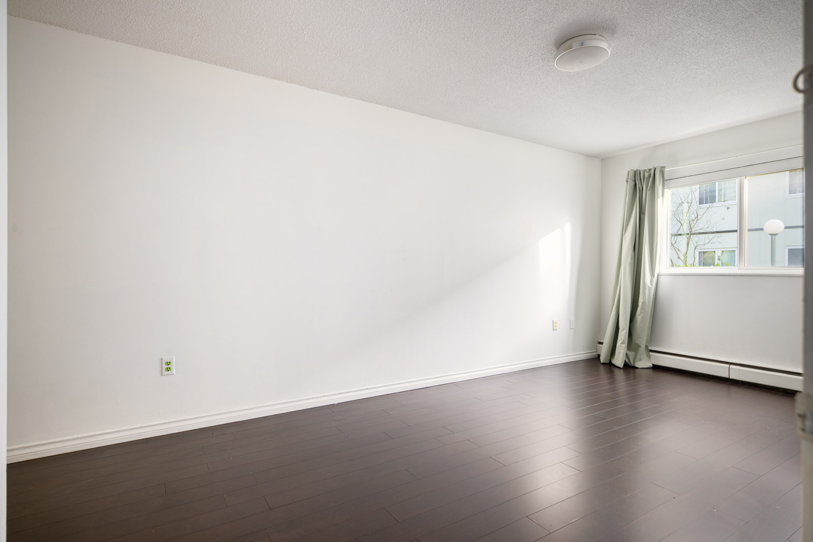 Empty room with white walls, dark wood flooring, a window with a single pale green curtain, and a round ceiling light.