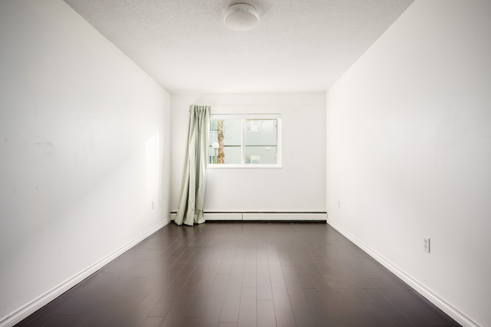 Empty white room with dark wood flooring, a window with light curtains, and a ceiling light fixture. Natural light enters from the window on the far wall.
