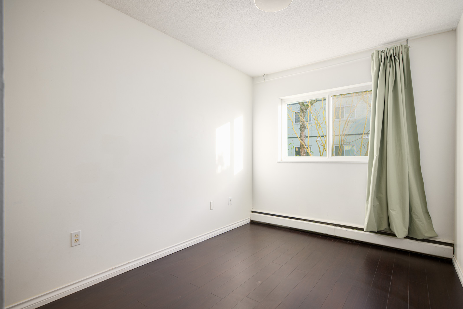 Empty room with white walls, dark wood floor, a window with light green curtains, and a baseboard heater beneath the window.