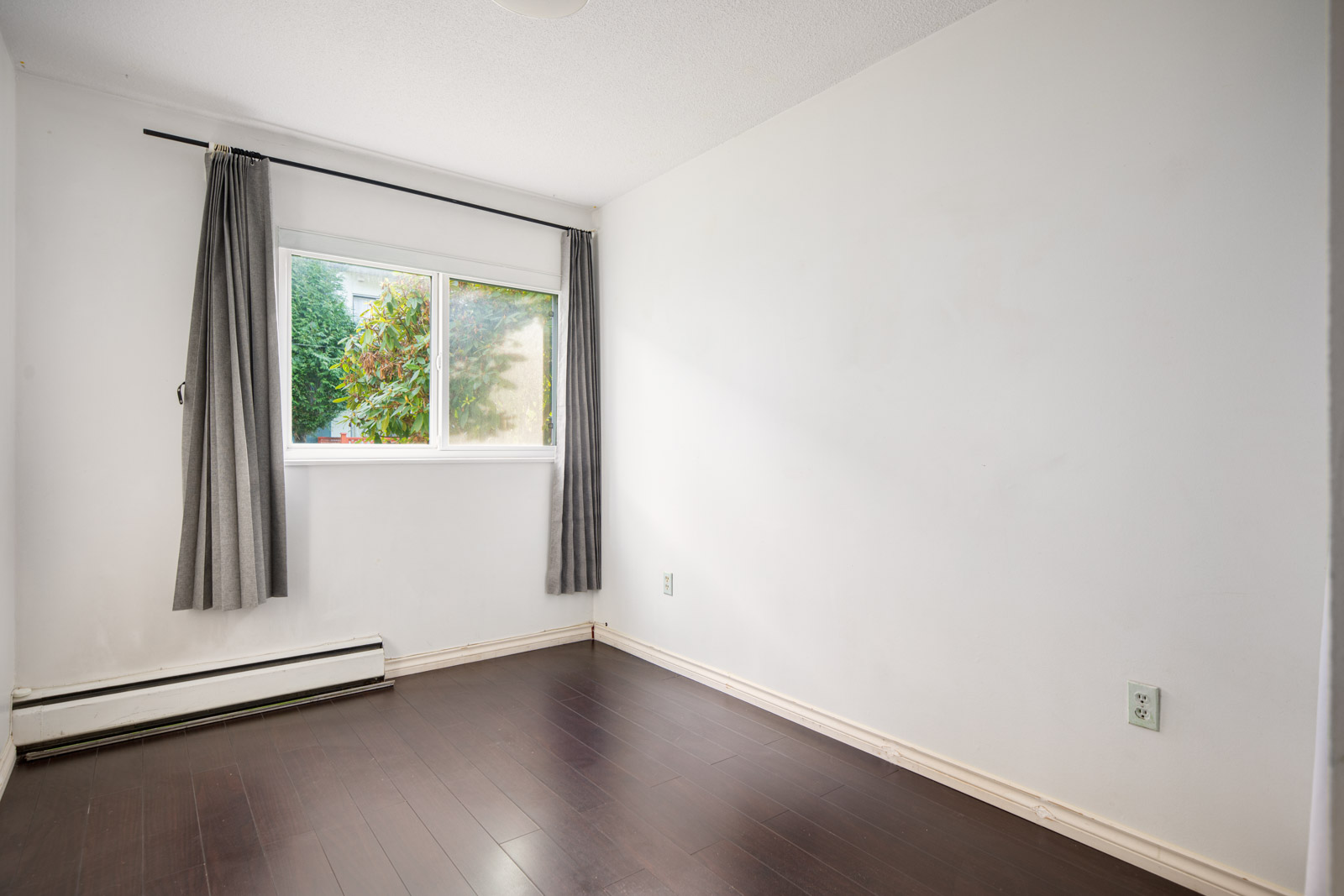 Empty room with white walls, dark wood flooring, a window with gray curtains, and a baseboard heater beneath the window.