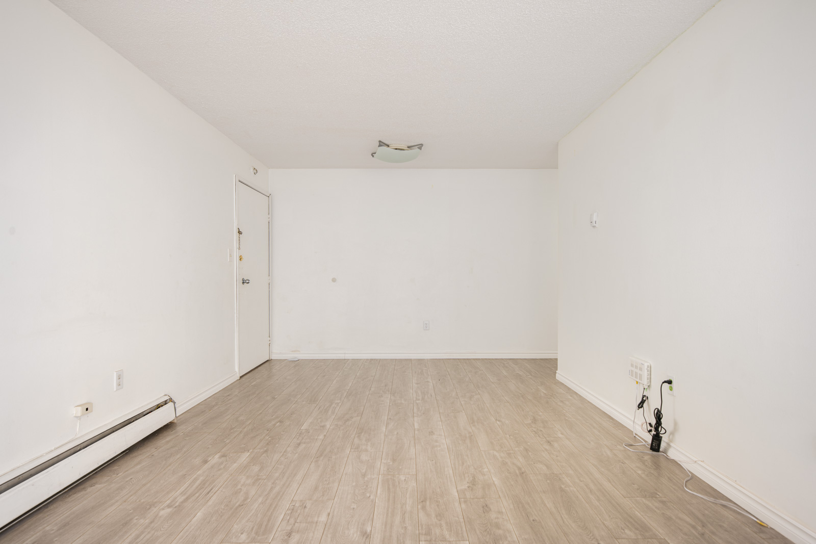 An empty, unfurnished room with white walls, wood-look flooring, a closed door, and exposed wall outlets and cables.