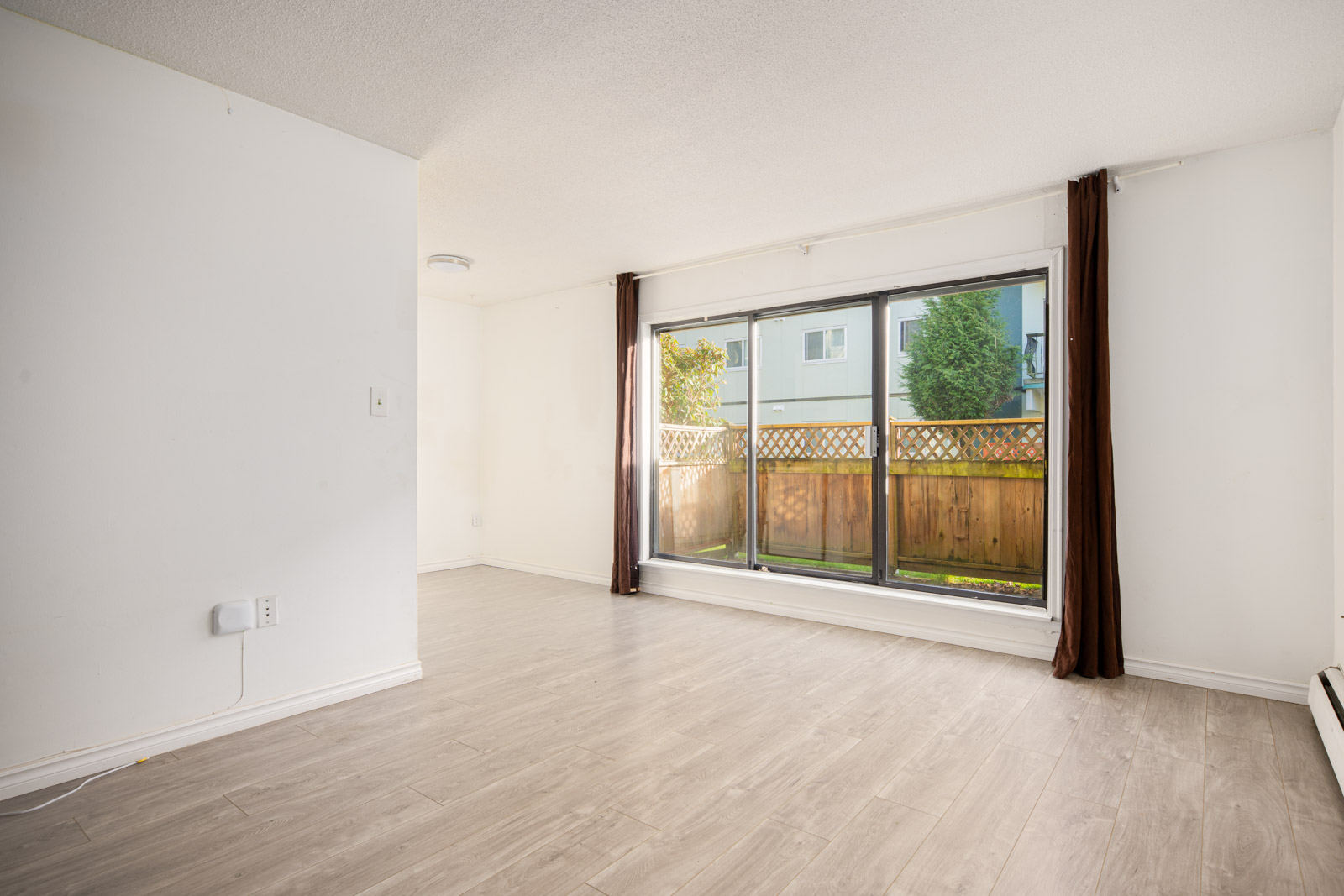 Unfurnished living room with light wood flooring, large window with brown curtains, and a view of a fenced outdoor area. White walls and ceiling, with a small nook visible to the left.