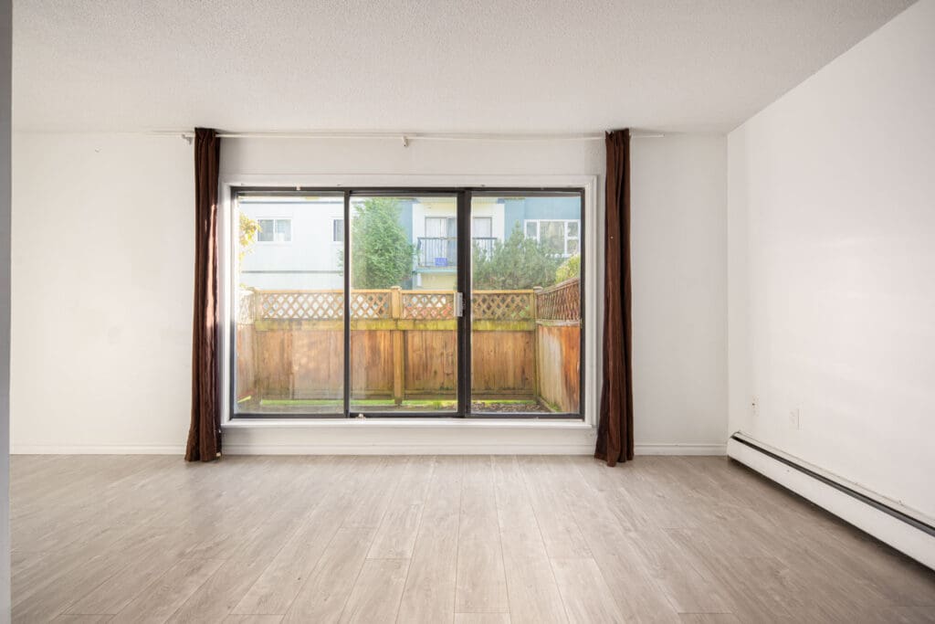 Empty room with light wood flooring, white walls, large window with brown curtains, and view of a fenced outdoor area.