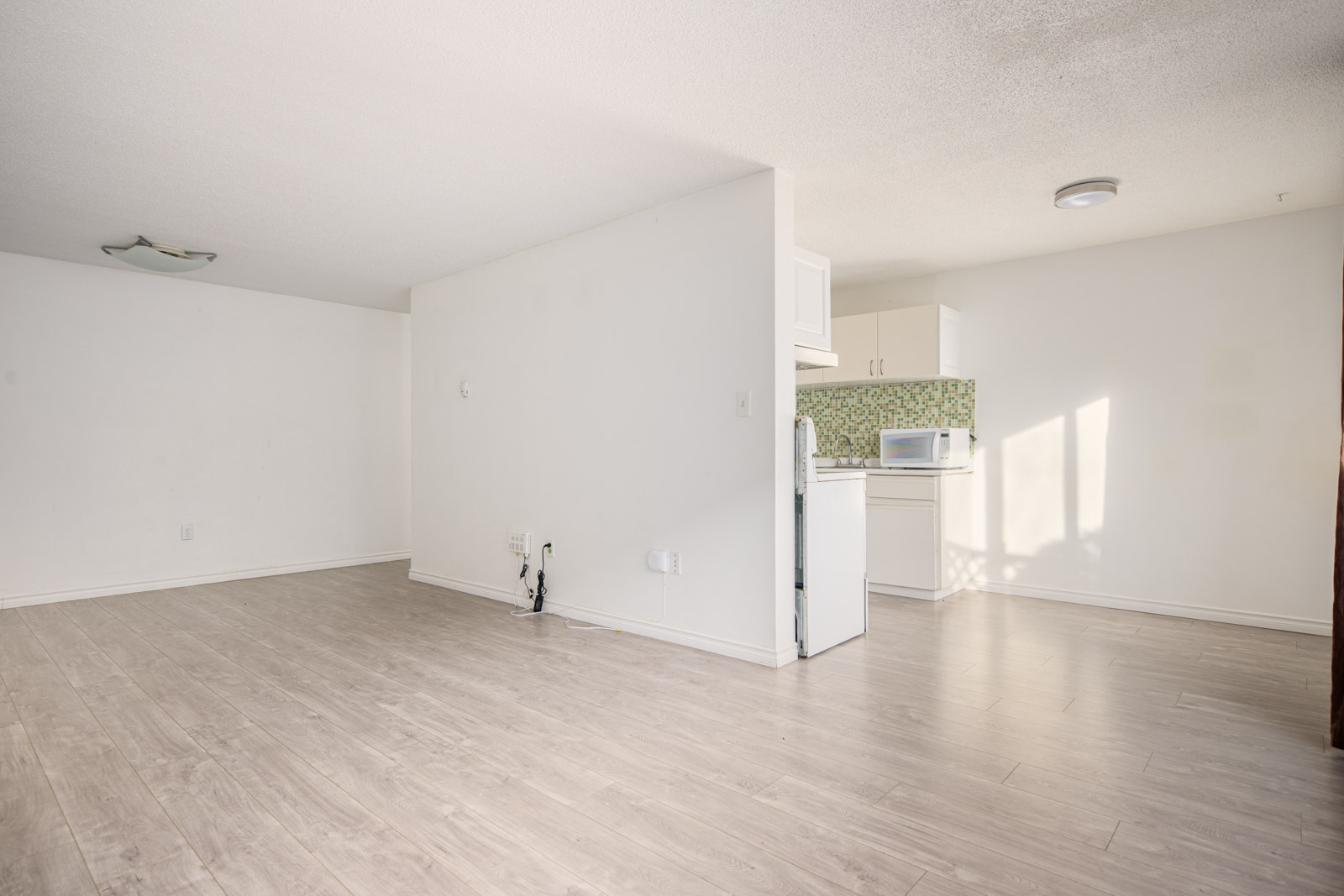 Empty room with light wood flooring, white walls, and a partial view of a small kitchen with green-tiled backsplash and a microwave on the counter.