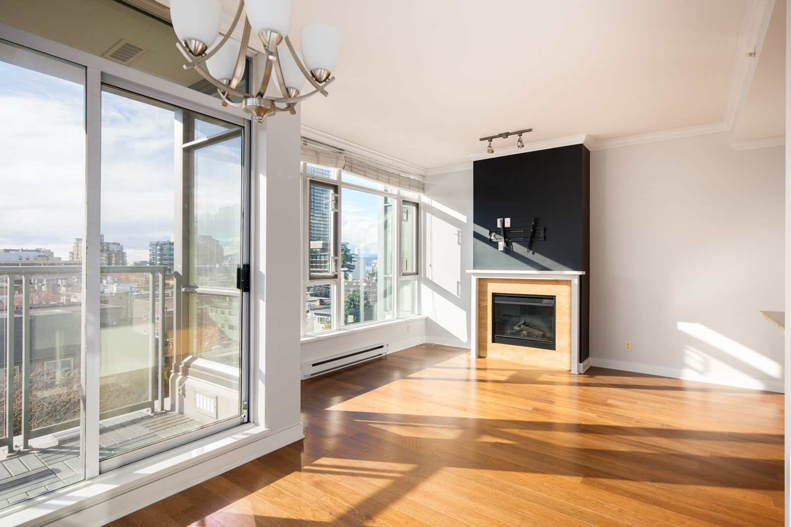 Bright, empty living room with hardwood floors, a fireplace, large windows, sliding glass door to a balcony, and city views. Modern chandelier hangs from the ceiling.