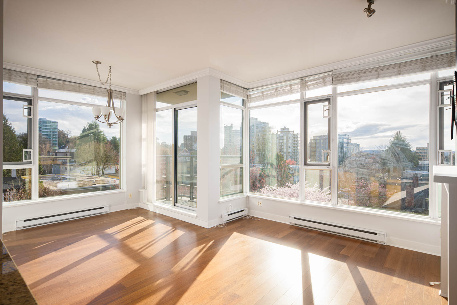 Bright, empty corner room with large windows, hardwood floors, and a cityscape view; sunlight casts shadows across the floor.