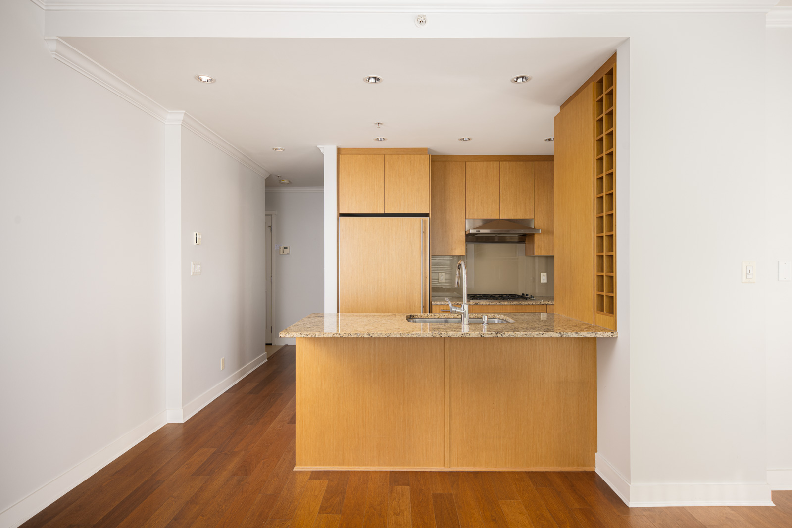 Modern kitchen with light wood cabinets, granite countertop island with sink, and stainless steel appliances, set against white walls and hardwood flooring.