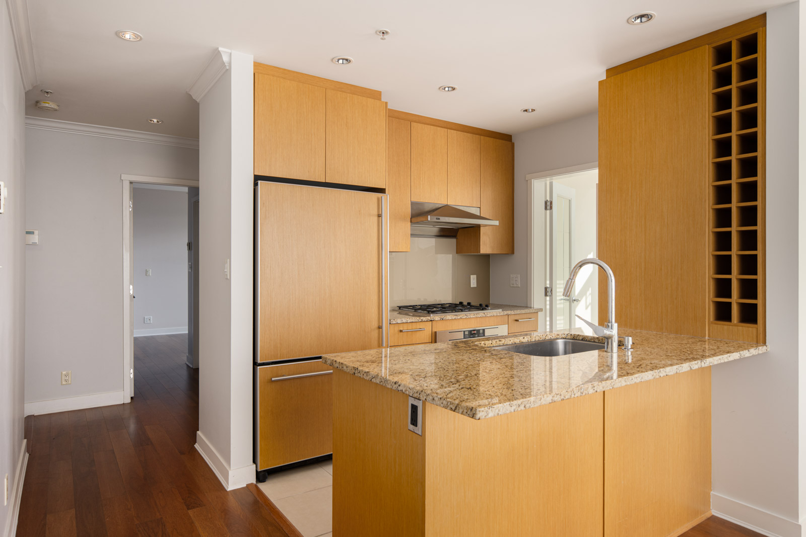 Modern kitchen with light wood cabinets, granite countertop island with sink, stainless steel appliances, and built-in wine rack. Hardwood floors and recessed lighting are visible.