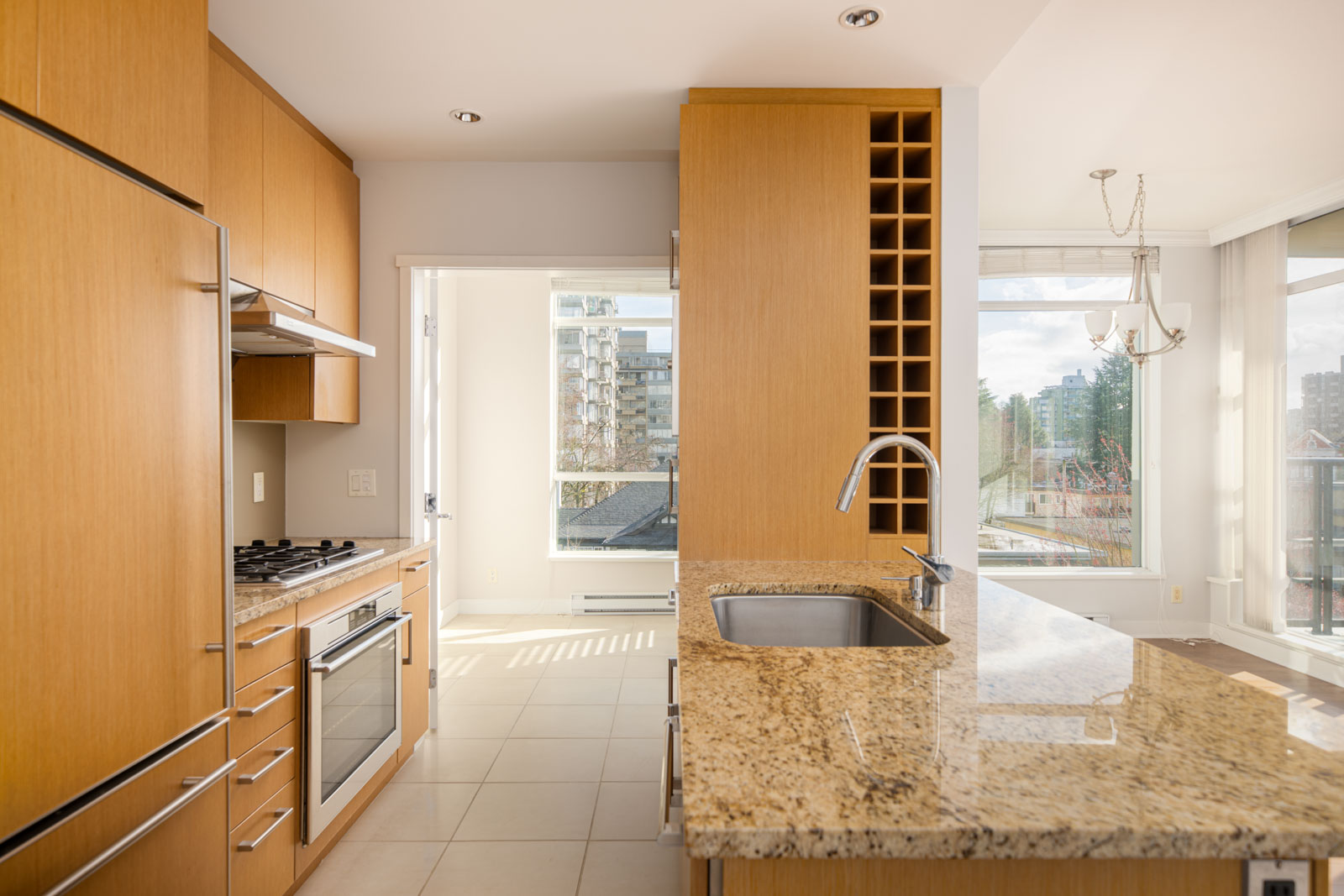 Modern kitchen with light wood cabinets, granite countertops, a built-in oven, gas stovetop, wine rack, and large windows letting in natural light.
