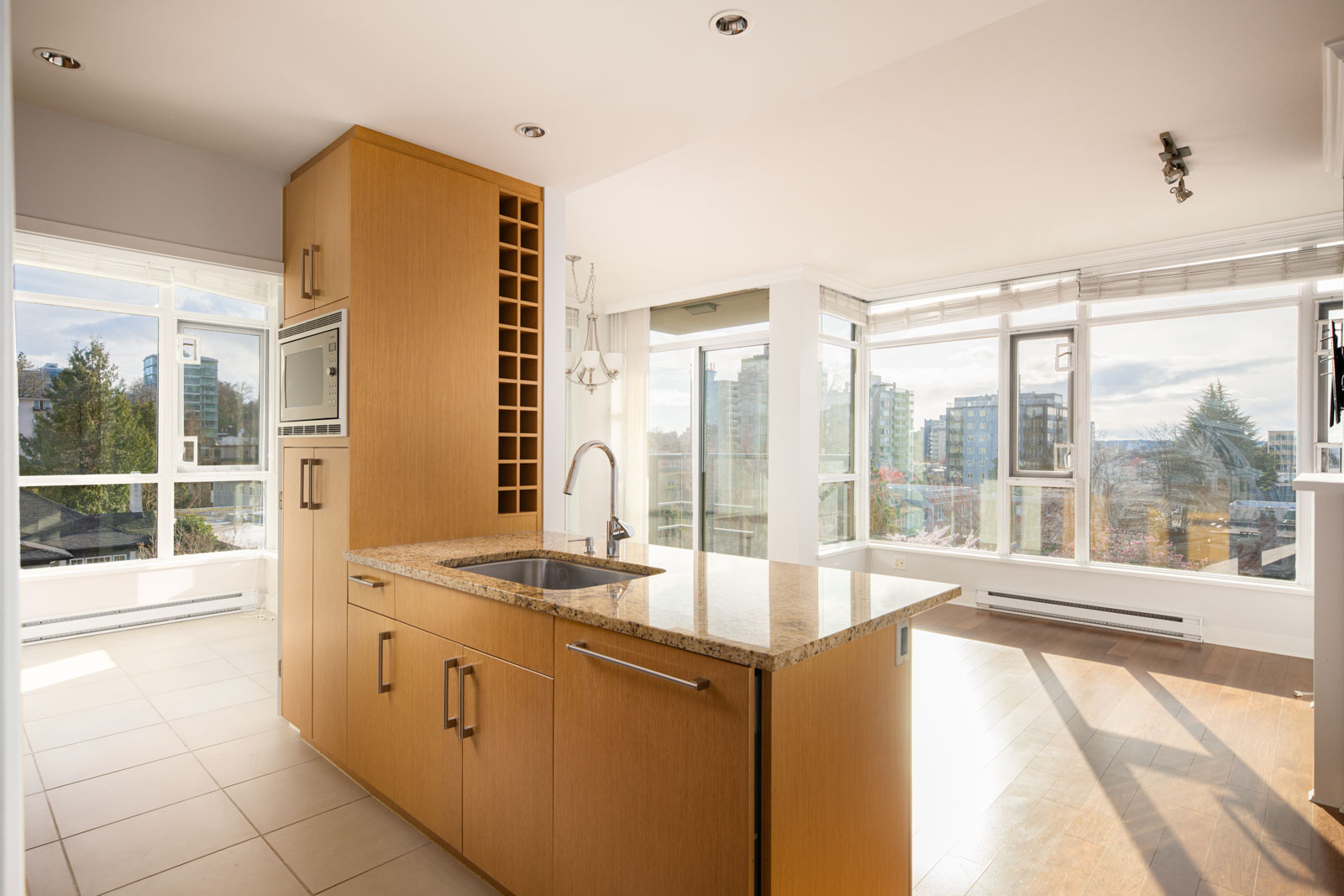 Modern kitchen with wooden cabinets, built-in microwave, wine rack, and island sink, set in a bright open-plan apartment with large windows and city views.
