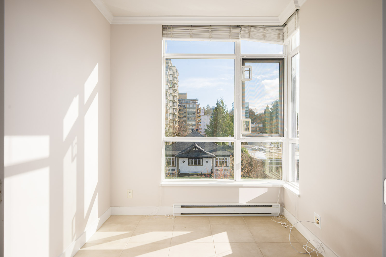Bright, empty room with light-colored walls and tiled floor, featuring a large window overlooking buildings, trees, and a partly cloudy sky.