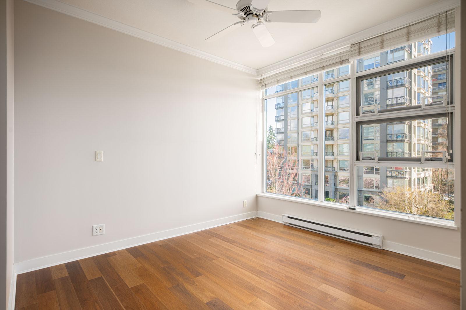 Empty room with hardwood floors, large windows, white walls, ceiling fan, and view of nearby apartment buildings through the windows.