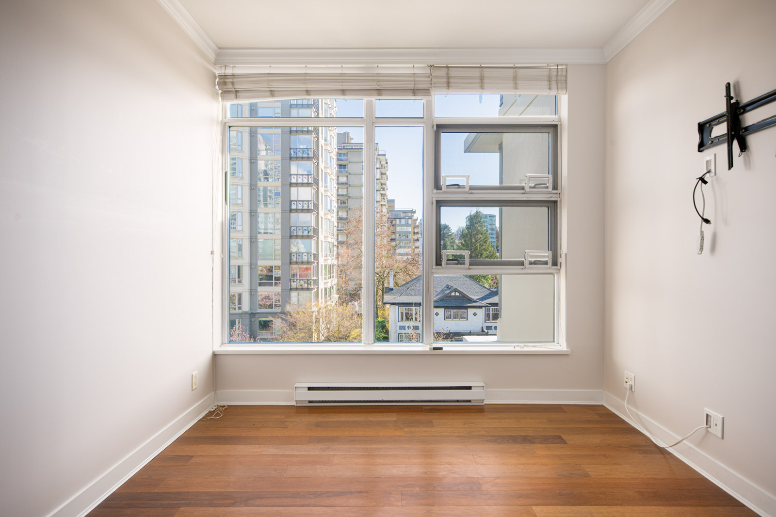 Empty room with light-colored walls, wood floor, large window overlooking city buildings, and a wall mount for a TV on the right.