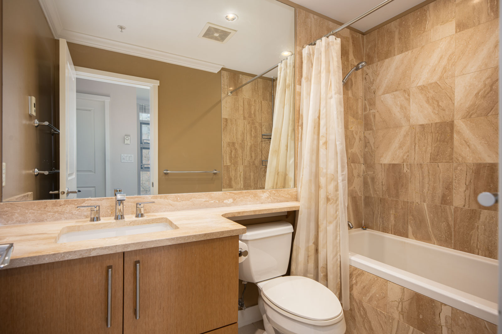 Modern bathroom with beige tile walls, a bathtub with a shower curtain, a toilet, a large mirror, and a vanity with a sink.