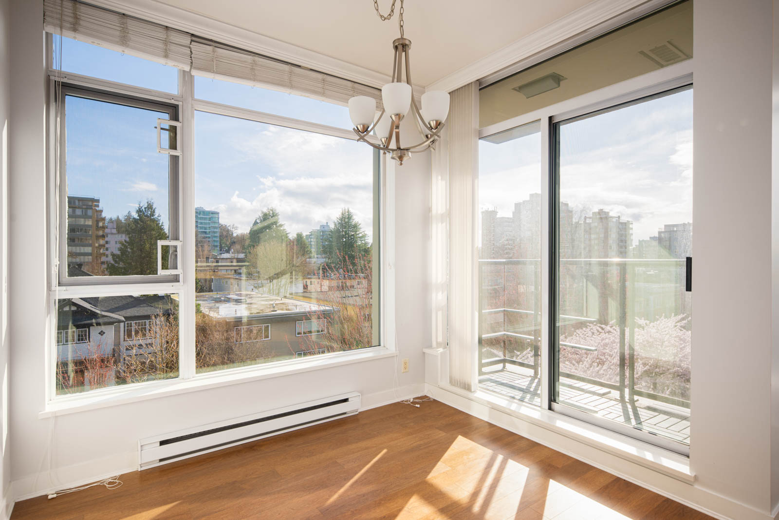 Bright, empty room with large windows, a glass door to a balcony, wooden floor, and a ceiling light fixture. City buildings and trees visible outside.