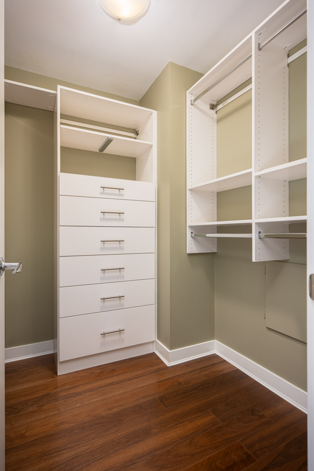 A walk-in closet with white built-in shelves, drawers, and hanging rods, featuring light green walls and a wood floor.