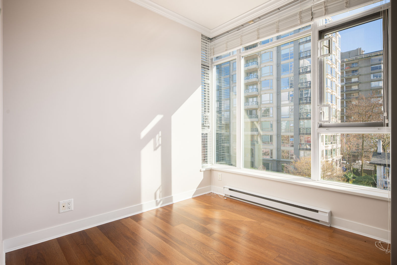 Empty corner of a sunlit room with large windows, hardwood floors, white walls, and a view of modern apartment buildings outside.