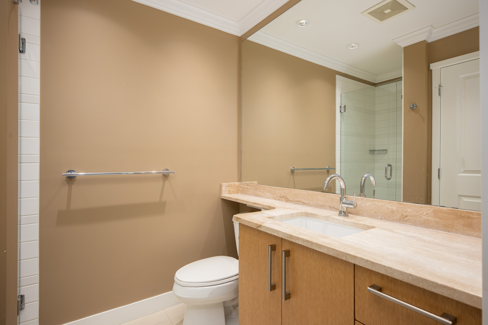 Modern bathroom with tan walls, a marble countertop, under-mount sink, large mirror, toilet, towel bar, and glass shower enclosure.