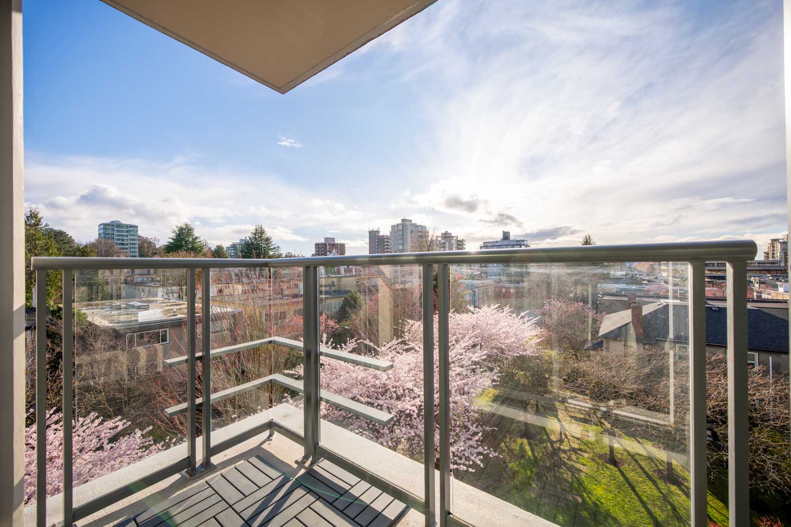 A modern balcony with glass railings overlooks a cityscape, trees with pink blossoms, and mid-rise buildings under a partly cloudy sky.