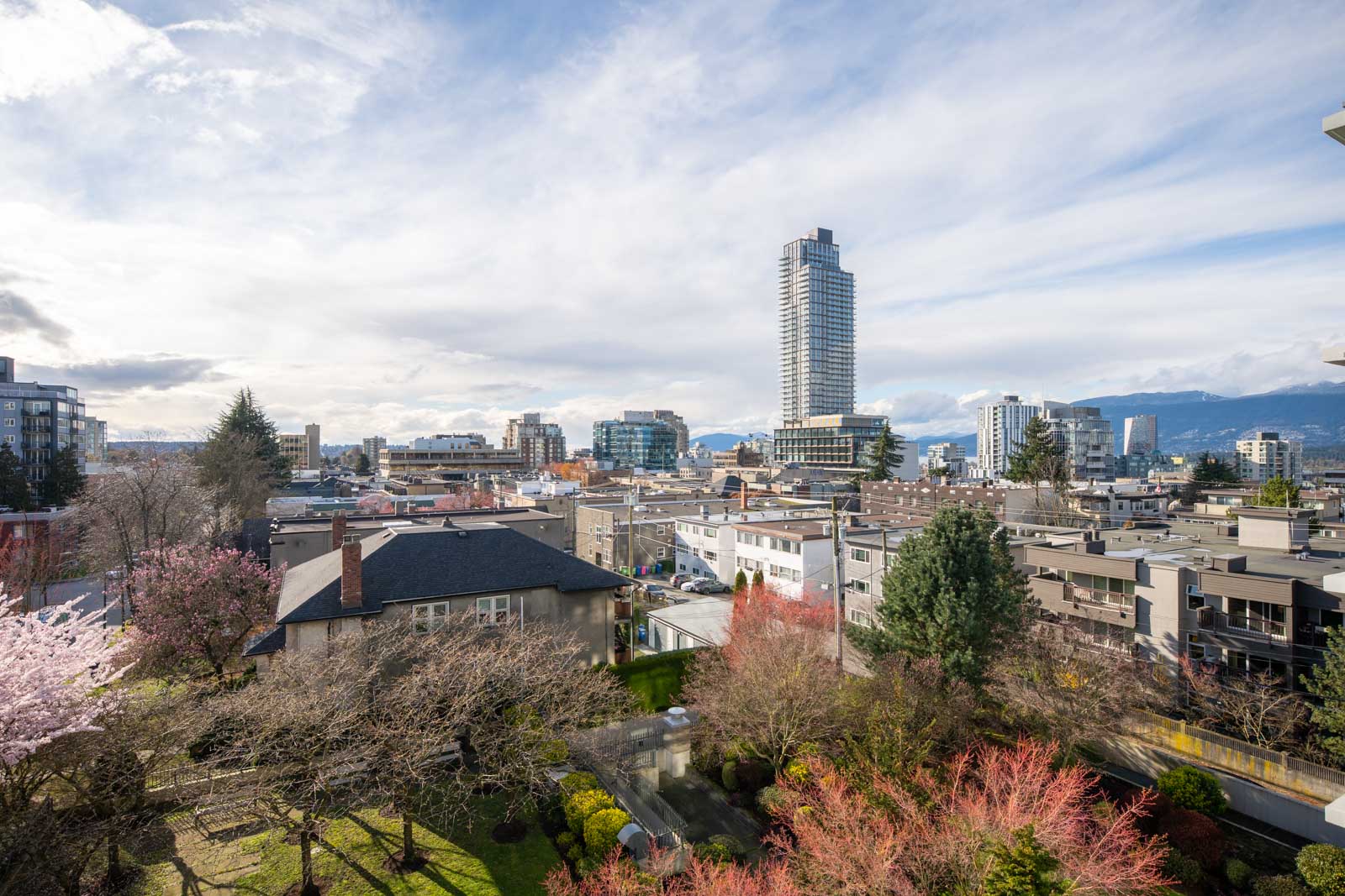 A cityscape featuring residential buildings, trees with sparse foliage, and a tall modern skyscraper under a partly cloudy sky.