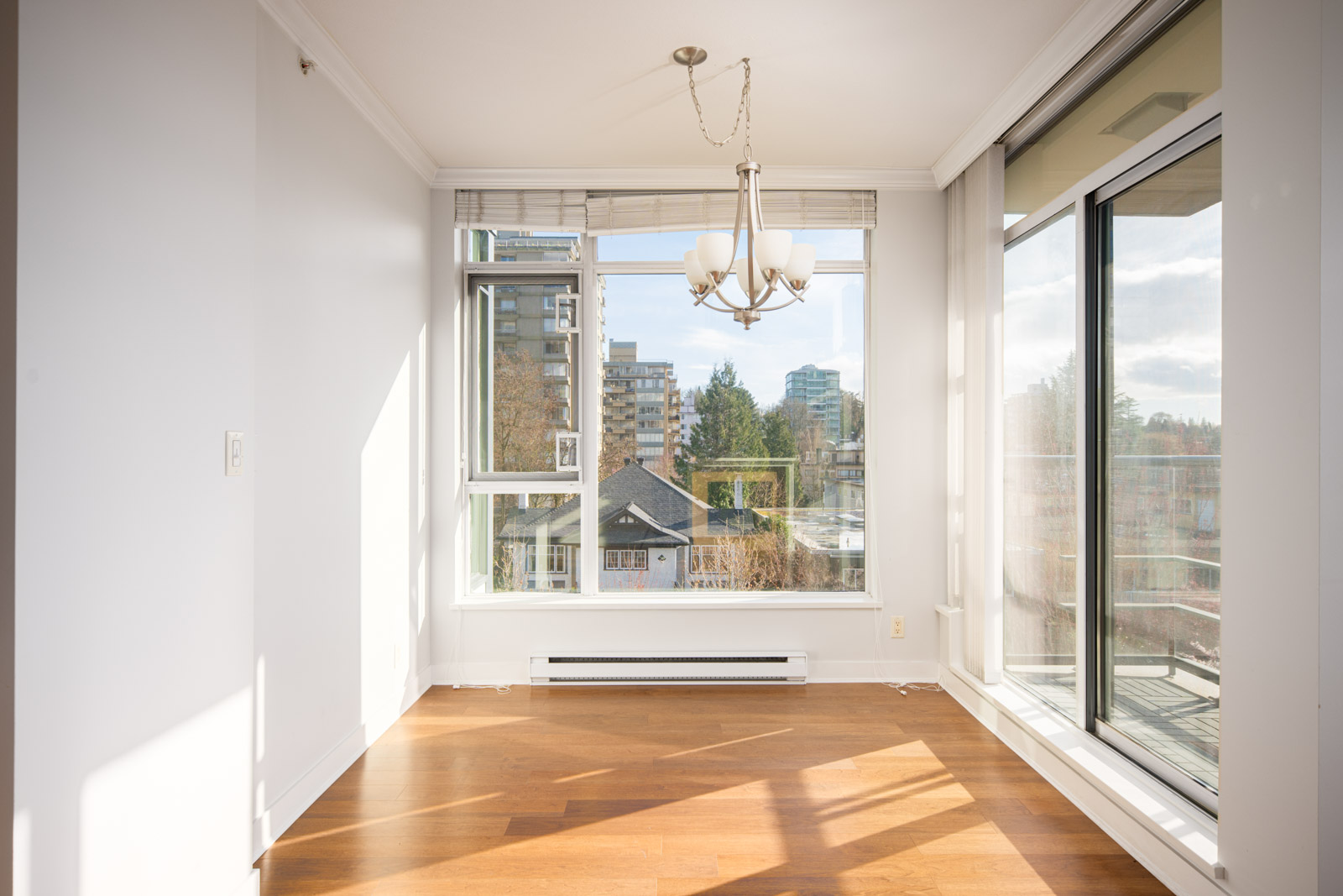Bright, empty room with wood flooring, a chandelier, large windows, and glass door opening to a balcony with a view of buildings and trees outside.