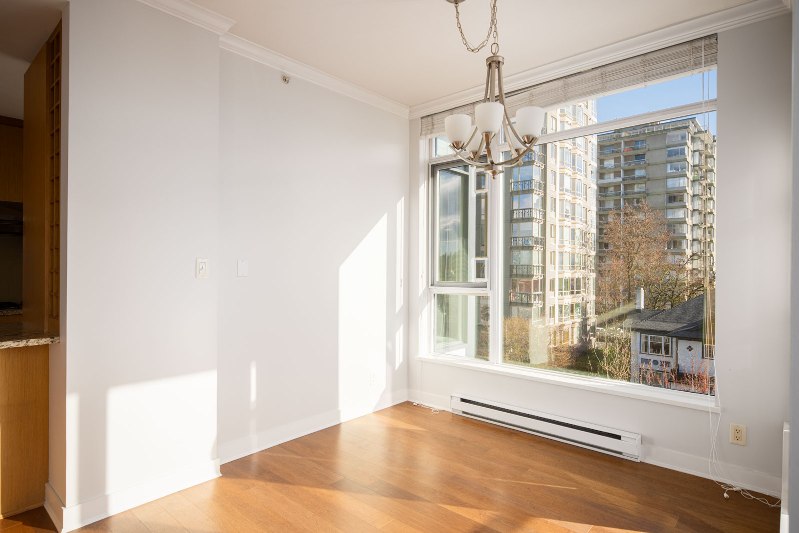 Bright, empty dining area with hardwood floors, a chandelier, large window, and a cityscape view of modern apartment buildings outside.