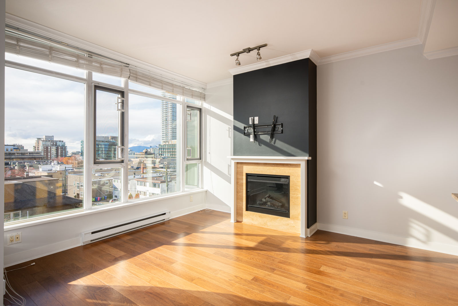 Bright, modern apartment living room with large windows, hardwood floors, a black accent wall, and a fireplace; cityscape view visible outside.