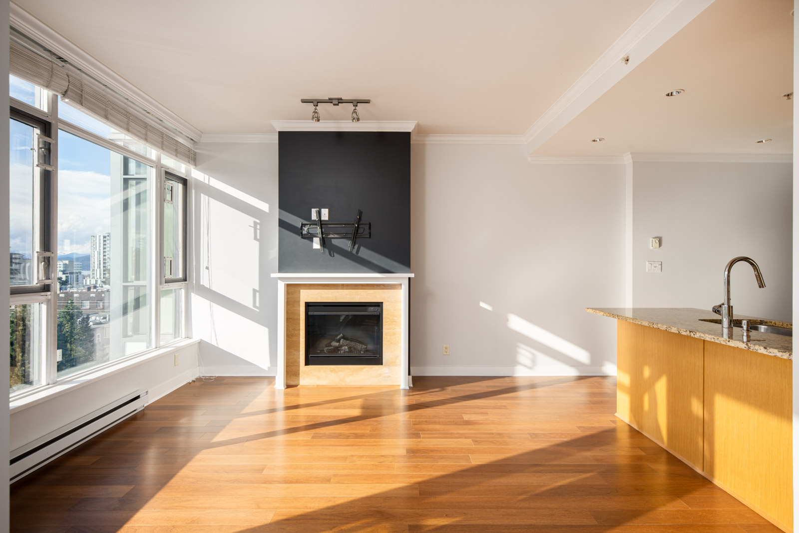 Bright, empty living area with large windows, hardwood floors, a fireplace with a black accent wall, and an open kitchen with a granite countertop.