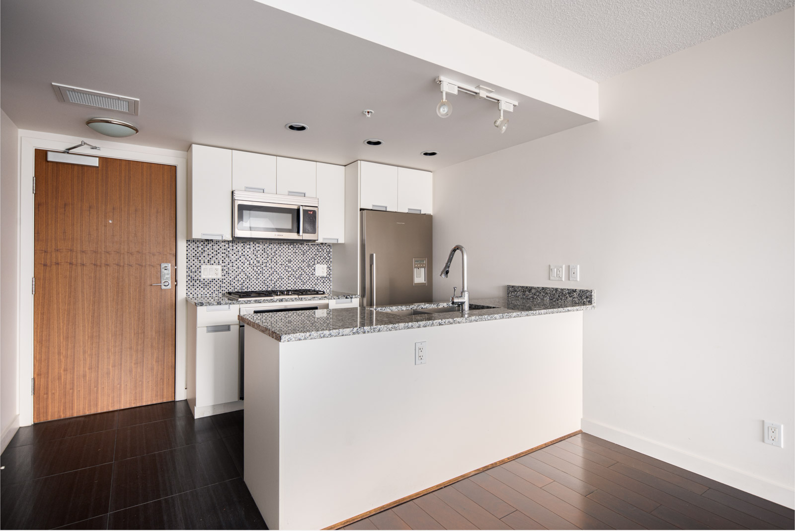 Modern kitchen with white cabinets, stainless steel appliances, granite countertops, a tile backsplash, and a wooden entry door.