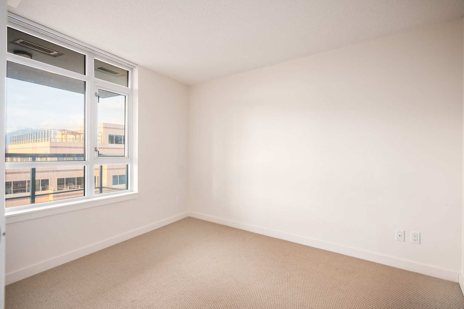 Empty room with beige carpet, white walls, and a large window showing a partial view of an adjacent building. Electrical outlets are visible on the lower right wall.